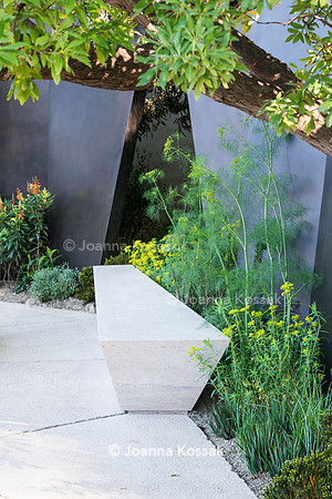 Seating area with  stone bench in front of irregular geometric bronze sculptures (fins) under tree Arbutus unedo