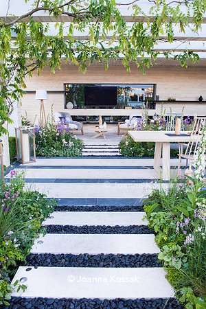 A path made from limestone paving slabs and separated with black oval stones leading to patio with wooden chairs and table (by Nils Verweij) surrounded by Stipa tenuissima, Tiarella 'Spring symphony',
