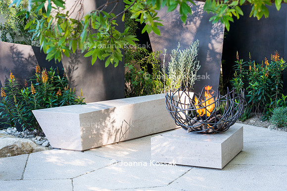Seating area with  stone bench and fire bowl in front of irregular geometric bronze sculptures (fins) under tree Arbutus unedo