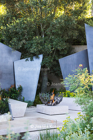 seating area with  stone benches and metal fire bowl in front of irregular geometric bronze sculptures (fins) surrounded by Quercus Ilex.